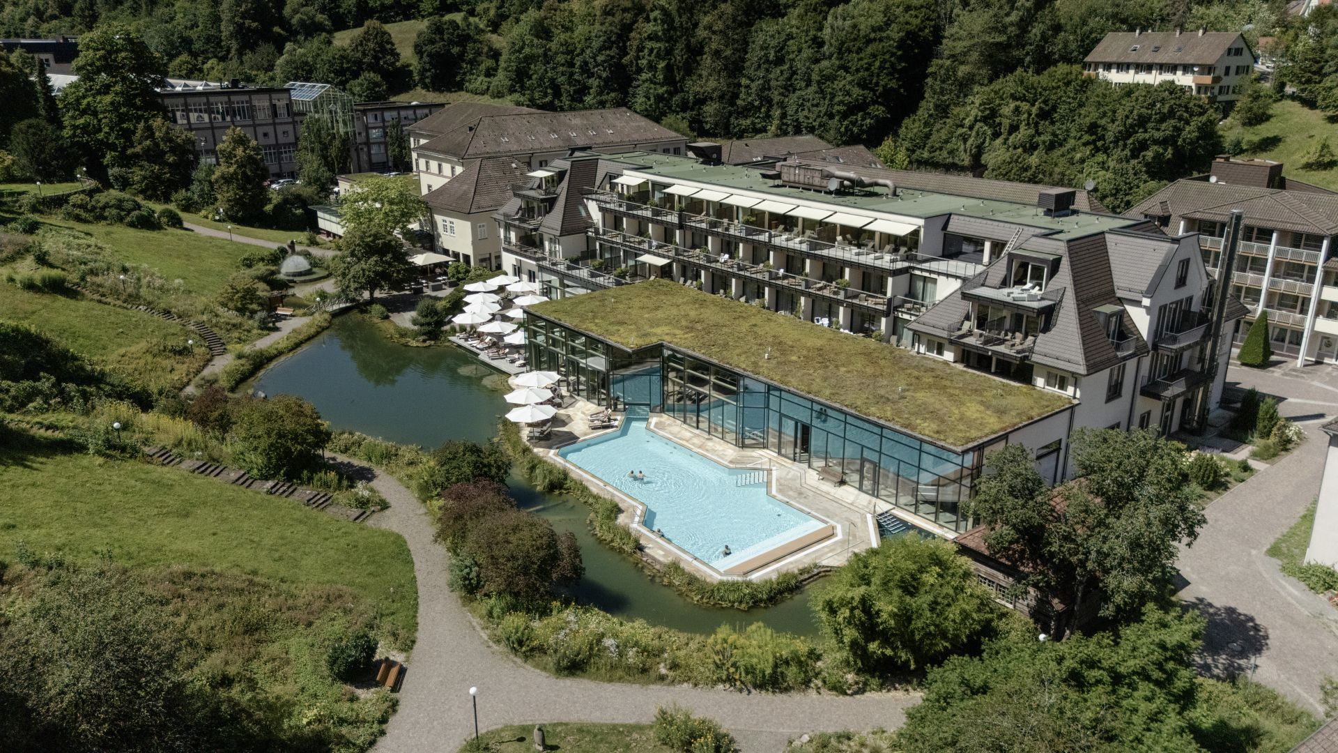 Das Hotel Therme Bad Teinach aus der Vogelperspektive. Neben den dazugehörigen Gebäuden ist ein Außenpool und eine bergige grüne Umgebung mit verschiedenen Bäumen und  Pflanzen zu sehen.