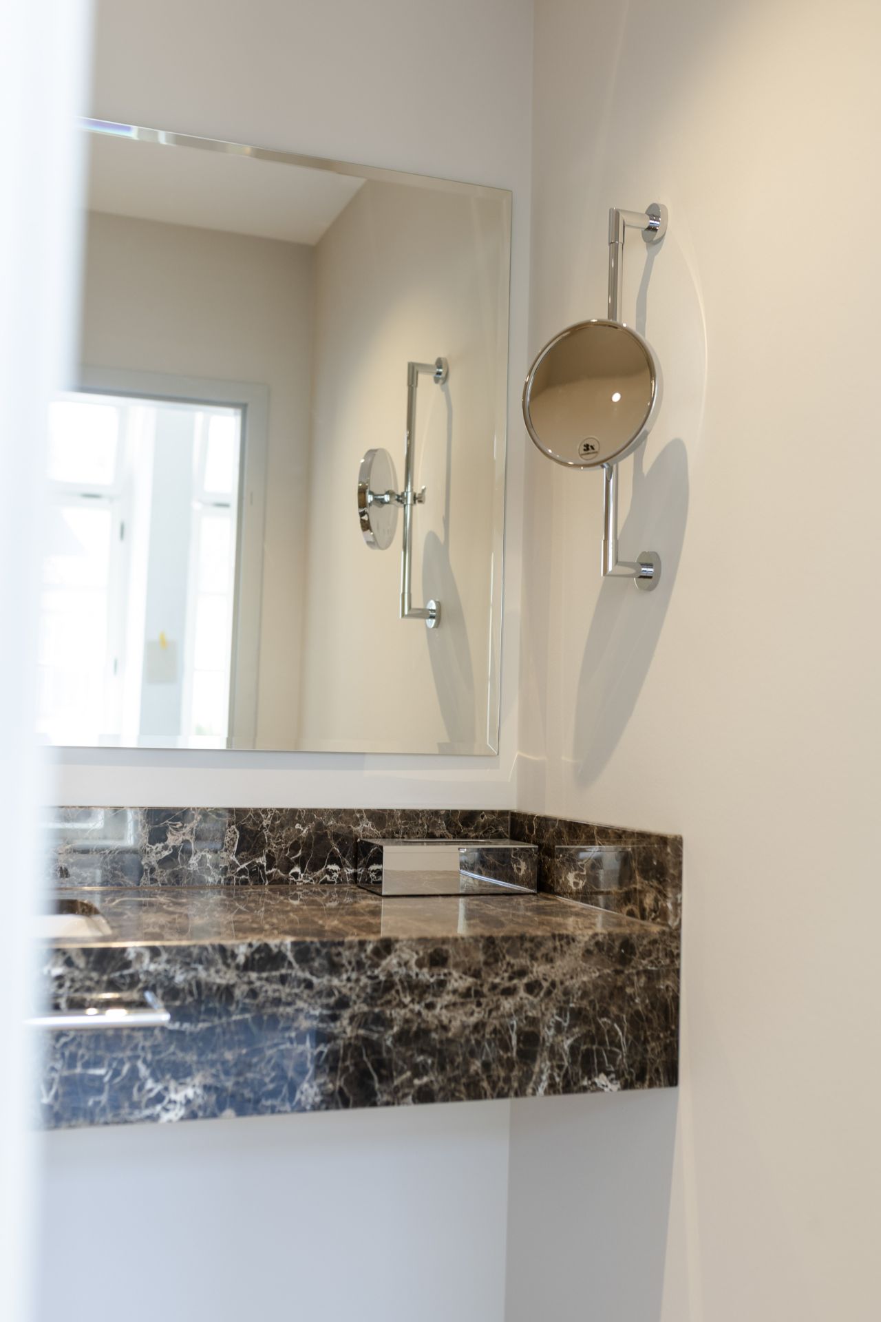 Detailansicht eines sanierten Hotelbads mit Wandspiegel, Wand-Kosmetikspiegel und Naturstein-Waschtisch.